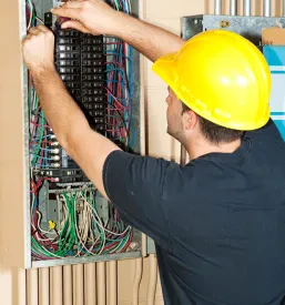 Električar v zaščitni čeladi in delovni obleki previdno menja varovalko v veliki industrijski razdelilni omarici.