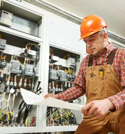 Inženir električar preučuje shemo pred nadzorno ploščo, kar je ključno za kvalitetne elektroinštalacije Nova Gorica, Goriška.