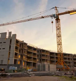 Panoramski pogled na gradbišče v jutranji zarji, z žerjavom in objekti v izgradnji pod svetlim nebom.