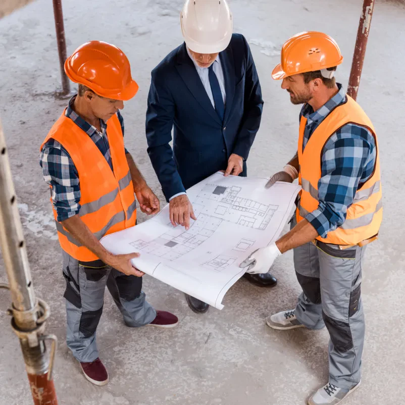 Pregled načrtov s strani gradbenikov Poslovnež in dva gradbenika v čeladah skupaj pregledujejo podrobne modre tlorise z višjega kota, kar poudarja timsko delo na projektu.