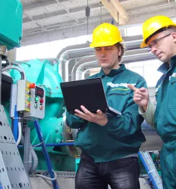 Dva industrijska delavca v zaščitni opremi sodelujeta pri pregledu načrtov na prenosniku, kar poudarja timsko delo pri kvalitetnem konstruiranju industrijskih strojev.