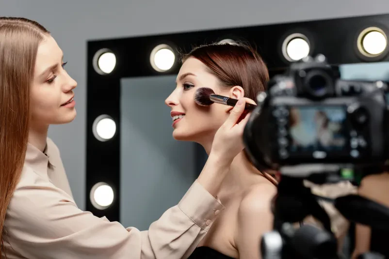 Vizažistka skrbno nanaša puder na model pred kamero Vizažistka z nasmehom nanaša puder na obraz lepega modela ob digitalni kameri, idealno za ličenje za fotografiranje v Ribnici.