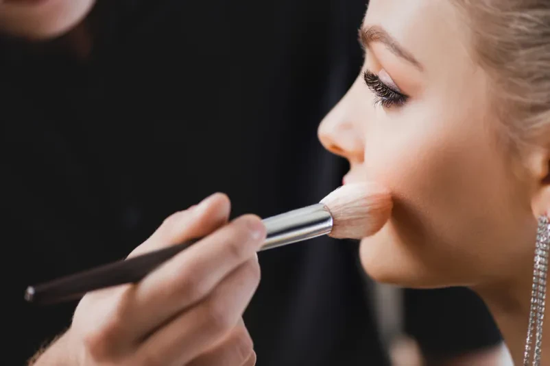 Vizažistka nanaša ličila na model v zakulisju Mlada vizažistka skrbno nanaša senčilo na veke privlačnega modela med pripravami v zakulisju, kar poudarja detajle ličenja.