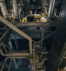Industrijski objekt s kovinskimi cevmi, prezračevalnimi kanali in jeklenimi nosilci. Večnivojska struktura poudarja kompleksnost okolja.