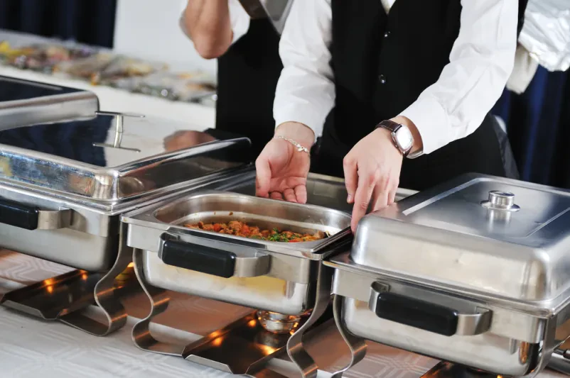 Skrbna priprava in aranžiranje jedi za bogat buffet Kuhar v beli uniformi skrbno aranžira izbrane jedi za catering buffet, poudarjajoč kakovost priprave hrane in vizualno privlačnost.