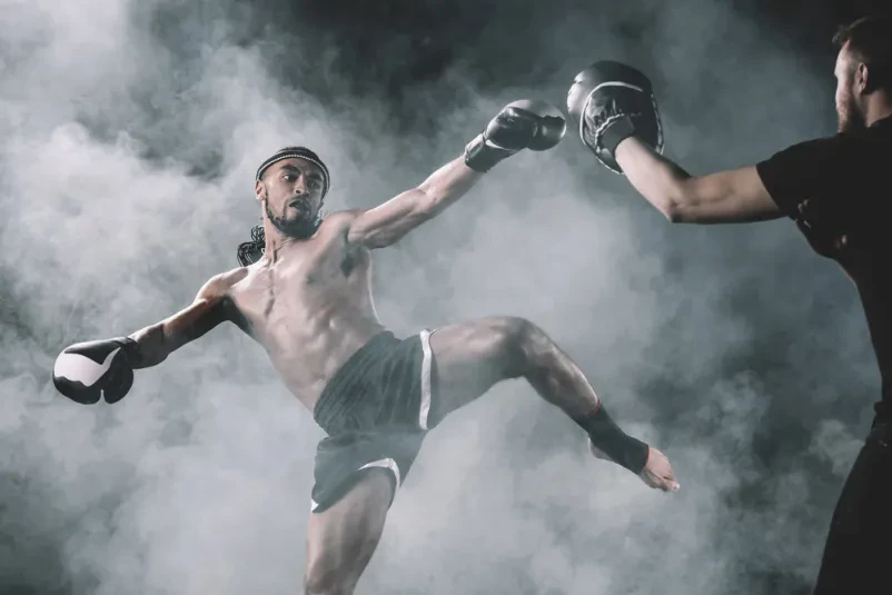 Osredotočen moški muay thai borec v gibanju, ki izvaja močan udarec. Silhueta na črnem ozadju poudarja njegovo moč in tehniko borilnih veščin.