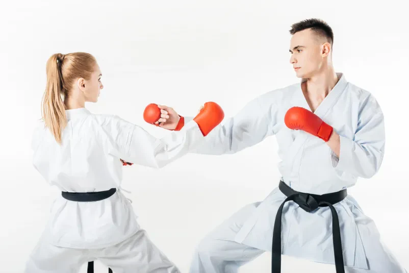 Moški in ženska karate borec z rdečim in črnim pasom med skupinskim treningom na beli podlagi. Izvajata tehniko bloka in napada v borilnih veščinah.