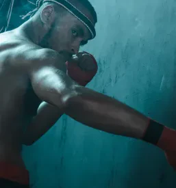 Pogled od strani na atletske roke muay thai borca z boksarskimi povoji, pripravljenega na intenziven trening tajskega boksa. Izraz na obrazu kaže na njegovo osredotočenost.