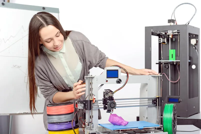 Mlado dekle s poudarkom spremlja delovanje sodobnega 3D tiskalnika v laboratoriju, kar prikazuje razvoj tehnologije.