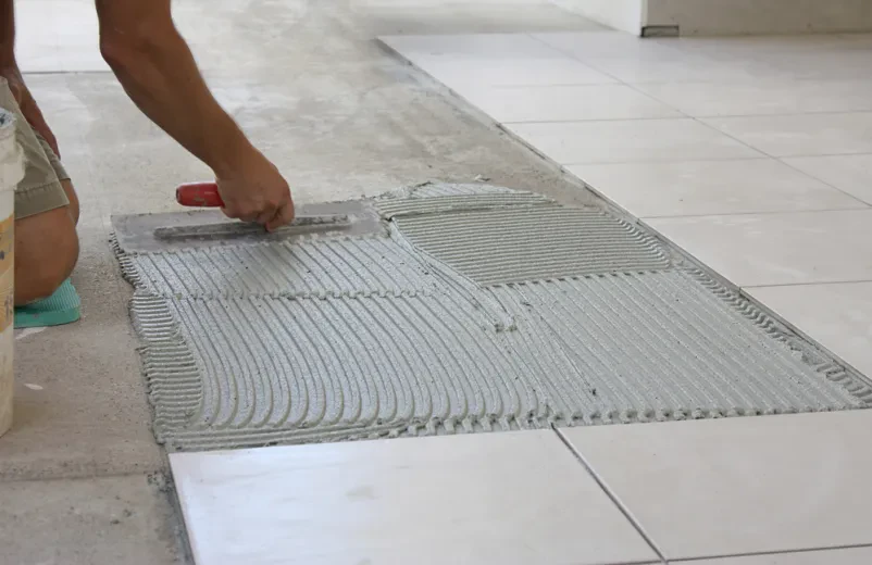 Mlad keramičar med delom na tleh Mlad moški keramičar kleči na tleh in natančno polaga nove svetle ploščice, kar kaže na predanost obrtnim delom. Njegovo delo prispeva k modernemu videzu prostora.