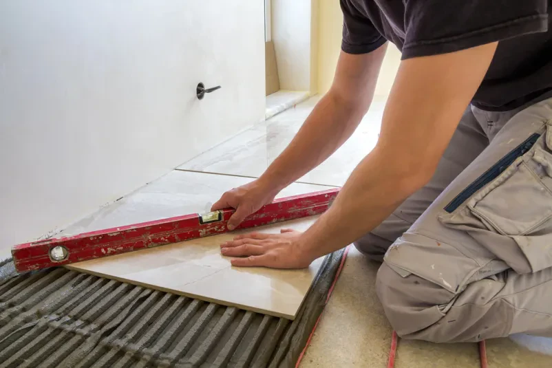 Polaganje keramike z električnim talnim gretjem Keramičar s pomočjo ročice namešča keramične ploščice na cementna tla, pod katerimi je rdeč električni sistem talnega gretja. To zagotavlja udobje in kakovostno Polaganje keramike Ajdovščina, Vipava, Nova Gorica.