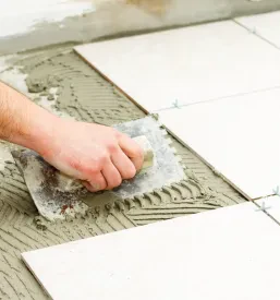 Delavec skrbno polaga svetle keramične ploščice na tla v kopalnici, natančno prilagaja vsak kos. Ta postopek zagotavlja trajnost in urejen videz površine.
