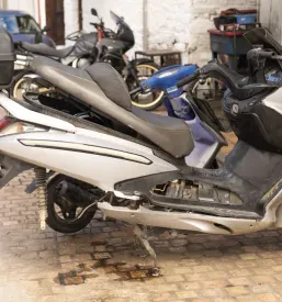 Razstavljen srebrn moped na stojalu pred delavnico, pripravljen na servis. V ozadju so vidna tudi druga motorna kolesa.