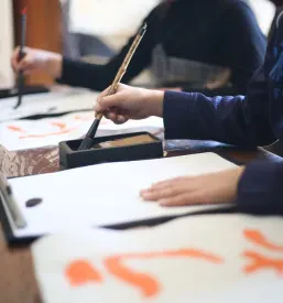 Udeleženec, verjetno otrok, se uči tradicionalne japonske kaligrafije s čopičem in črnilom, kar je odlična priložnost za učenje japonske kulture.
