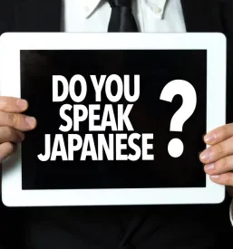 Poslovnež v obleki drži tablico, na kateri je vprašanje 'Do You Speak Japanese?', kar poudarja pomembnost jezikovnih veščin v poslovnem svetu.