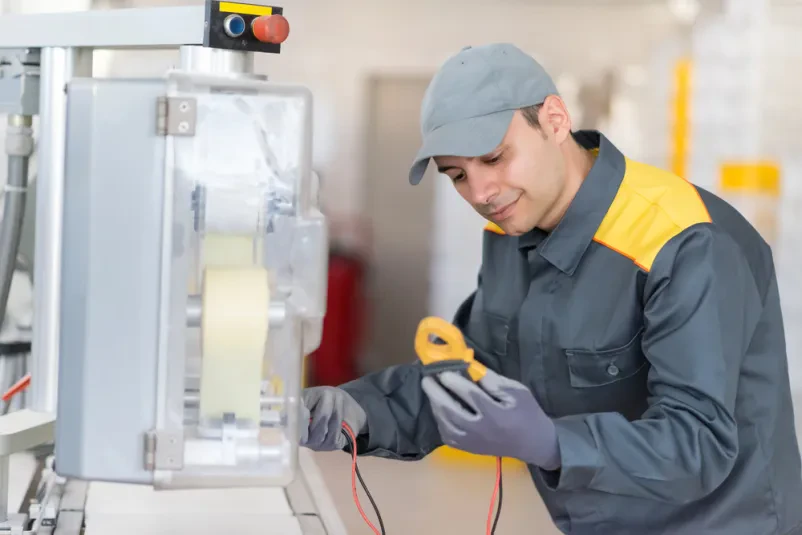 Industrijski električar pri popravilu stroja Industrijski električar v delovni obleki in z orodjem v roki predano popravlja industrijski stroj, s poudarkom na natančnosti in strokovnosti pri vzdrževalnih delih.