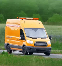 Beli servisni kombi z lestvijo na strehi vozi po podeželski cesti, kar ponazarja mobilnost in dostopnost Servisa na terenu po Sloveniji za najrazličnejše elektronske stroje in naprave.