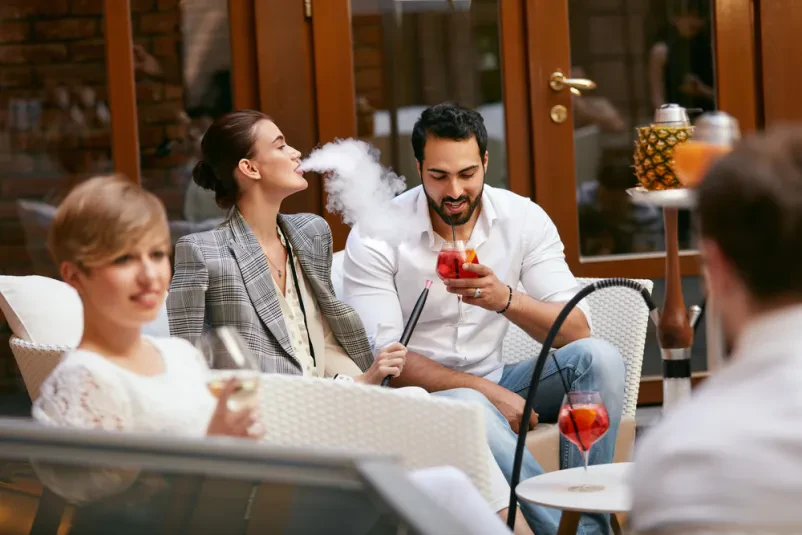 Gostje uživajo v šishi in koktajlih v baru Eleganten par kadi šišo in uživa v osvežilnih koktajlih v trendovskem lokalu, obdan z belim pohištvom in sproščeno atmosfero.