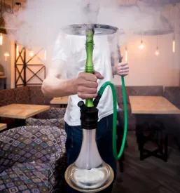 Mlad moški natakar skrbno pripravlja nargilo z ogljem in okusnim tobakom, kar ustvarja avtentično doživetje v trendovskem shisha baru.