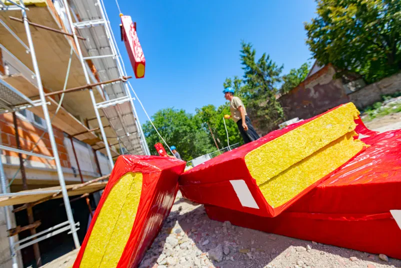 Velik kup toplotnoizolacijskega materiala, ovitega v folijo, na gradbišču. Delavec s pomočjo dvigala namešča en paket kamene volne na kavelj, da ga dvigne na fasado in izolira stanovanjsko stavbo.