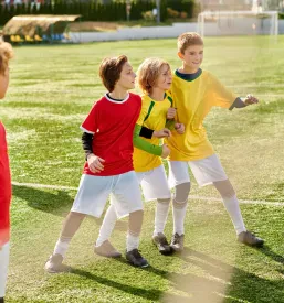 Vesela skupina mladih nogometašev zmagoslavno stoji na nogometnem igrišču, združena v veselju in tovarištvu, kar je bistvo vsakega športnega društva.