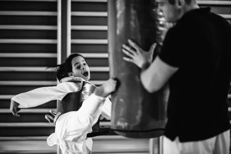 Mlada deklica trenira udarce na vreči v taekwondo šoli Deklica v belem taekwondo kimonu močno brca boksarsko vrečo. Trening tekwando v Ljubljani, v Jaršah, krepi moč in izboljšuje tehniko.