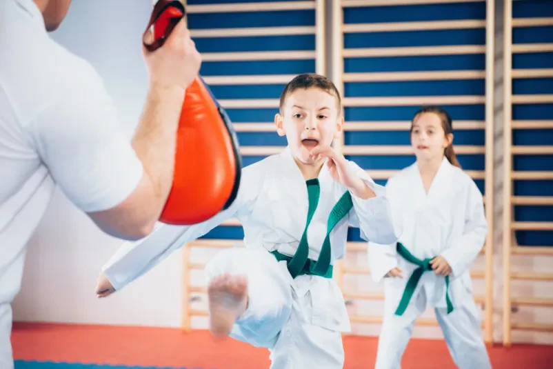 Skupina otrok na treningu taekwondoja s trenerjem Skupina otrok v taekwondo uniformah pozorno posluša trenerja. Gre za prikaz skupinskega treninga borilnih veščin v športnem klubu.