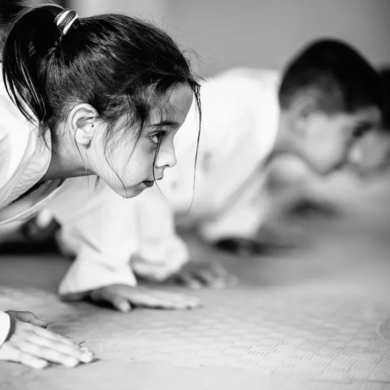 Mlada deklica na intenzivnem treningu taekwondoja Mlada deklica zbrana izvaja gibe taekwondoja v uniformi (doboku) med treningom borilnih veščin, poudarjajoč fokus in disciplino.