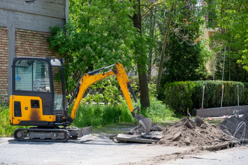 Rumeni mini bager koplje jarek ob cesti Rumeni mini bager koplje dolg jarek ob tlakovani cesti, medtem ko v ozadju stoji zidana stavba, obdana z zelenimi drevesi.