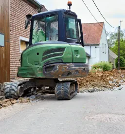 Zeleni mini bager stoji na ozki podeželski cesti poleg kupa izkopane zemlje, v daljavi pa so vidne hiše in drevesa.