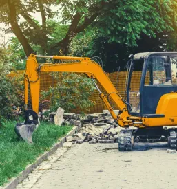 Majhen bager se nahaja na pločniku v mestu, blizu zelenice, kar nakazuje na potek manjših komunalnih ali vzdrževalnih del.