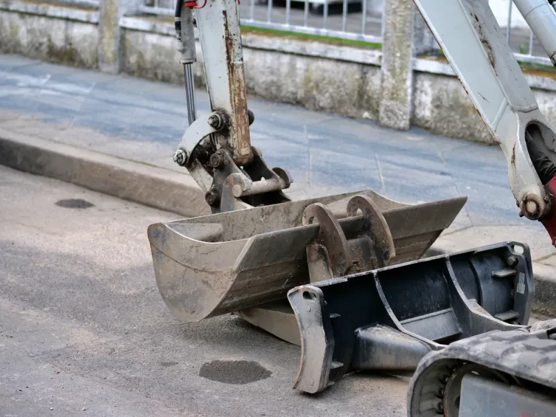 Navezave mini bagra na asfaltni površini Od blizu je prikazana robustna priključna oprema mini bagra na asfaltu, z obrabljeno žlico in črnim rezilom. Ugoden servis mini bagrov Goriška, Slovenija zagotavlja zanesljivo delovanje.