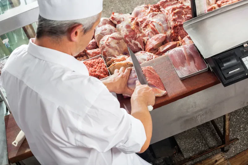 Mesar reže meso v prodajni vitrini Mesar skrbno reže velik kos mesa v hladilni vitrini mesnice, kar omogoča strankam vpogled v svežo ponudbo in predelavo mesa.