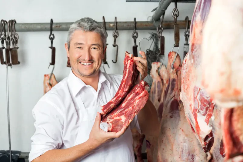 Mesar s svežim mesom v klavnici Zreli mesar z nasmehom ponosno drži velik kos svežega rdečega mesa v klavnici, kar kaže na kakovosten Zakol živine Savinjska, Šaleška.
