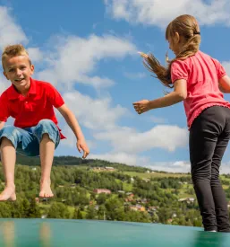 Dva skandinavska otroka, fant in deklica, se zabavata in veselo skačeta na velikem napihljivem trampolinu pod modrim nebom.