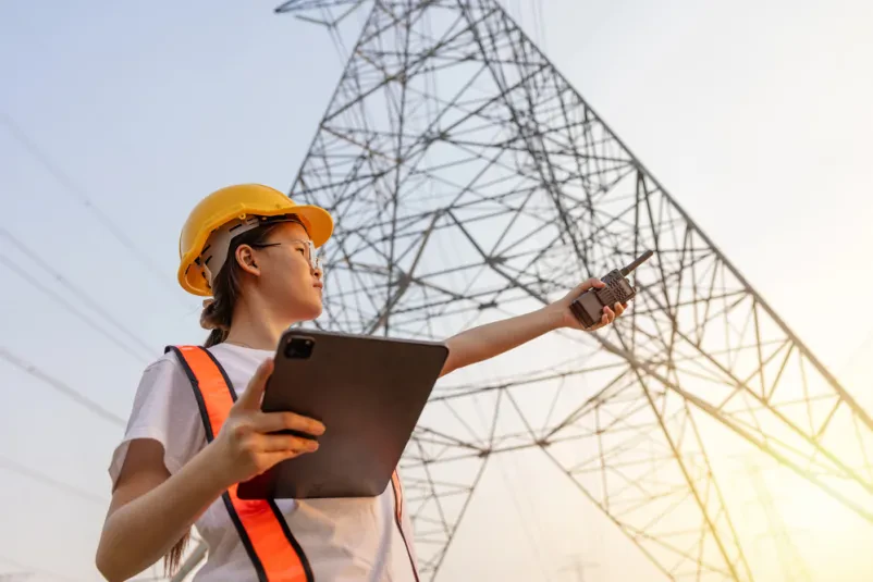 Inženirka pri pregledu daljnovodnega omrežja Inženirka s čelado in beležnico pregleda daljnovod, kar poudarja pomen zanesljive dobave električne energije v Ljubljani in širše.