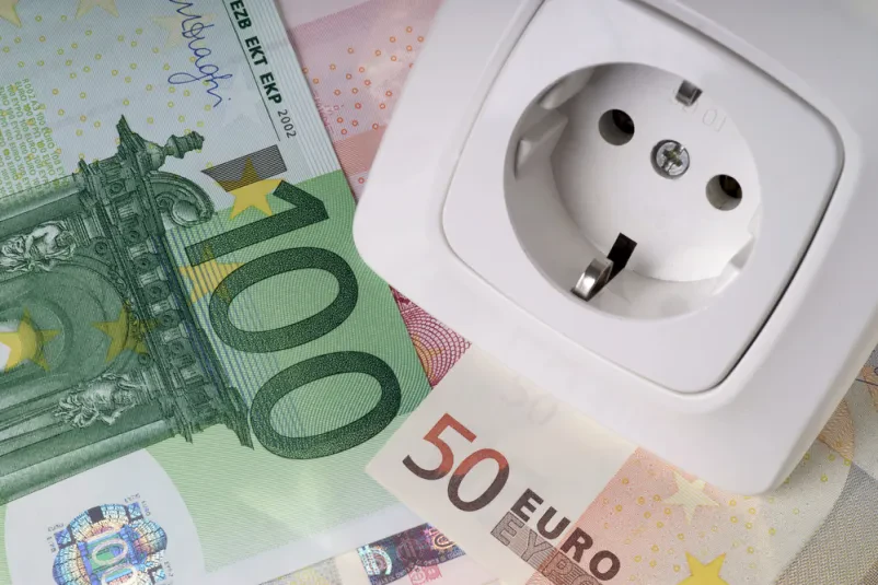 Cena električne energije in finančni vidiki Prikaz evrskih bankovcev, ovitih okoli električne vtičnice, simbolizira stroške in ceno električne energije za gospodinjstva in podjetja.