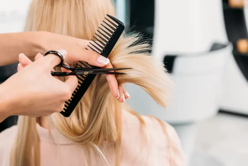 Friziranje blond las Stilistka skrbno striže konice blond las mlade ženske v frizerskem salonu, poudarja nego las in natančnost.