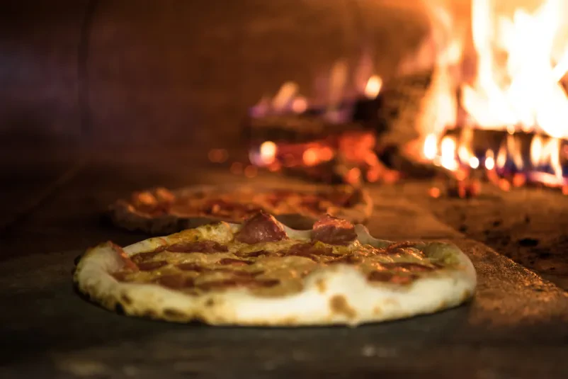 Približan pogled na okusno italijansko pico, ki se peče v tradicionalni krušni peči, z živahnimi plameni v ozadju, kar zagotavlja hrustljavo skorjo.