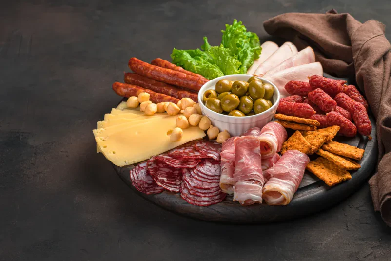 Okrožen pladenj s sirom in mesninami Razkošen okrogel servirni pladenj, obložen z različnimi narezanimi mesninami in siri, okrašenimi s svežimi zelišči in olivami, postavljen na rjavem ozadju.