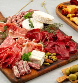 Obložena miza ponuja raznolike antipaste in prigrizke, vključno z narezano klobaso, pršutom, sirom in krekerji, ob kozarcih rdečega vina.