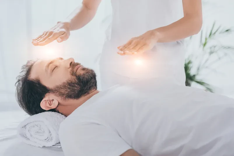 Moški z zaprtimi očmi leži sproščen, medtem ko prejema reiki zdravljenje. Fokus je na njegovem notranjem miru.