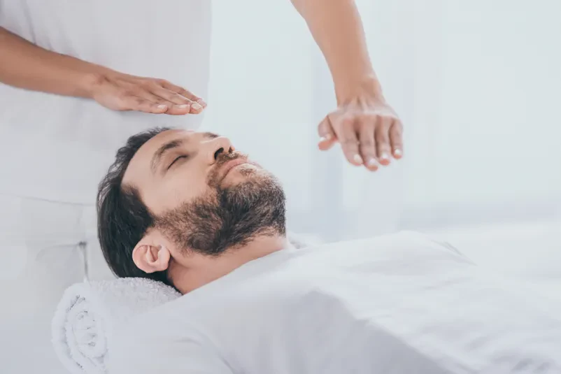 Spokojen bradati moški leži na masažni mizi, medtem ko terapevt izvaja reiki seanso s položenimi rokami.