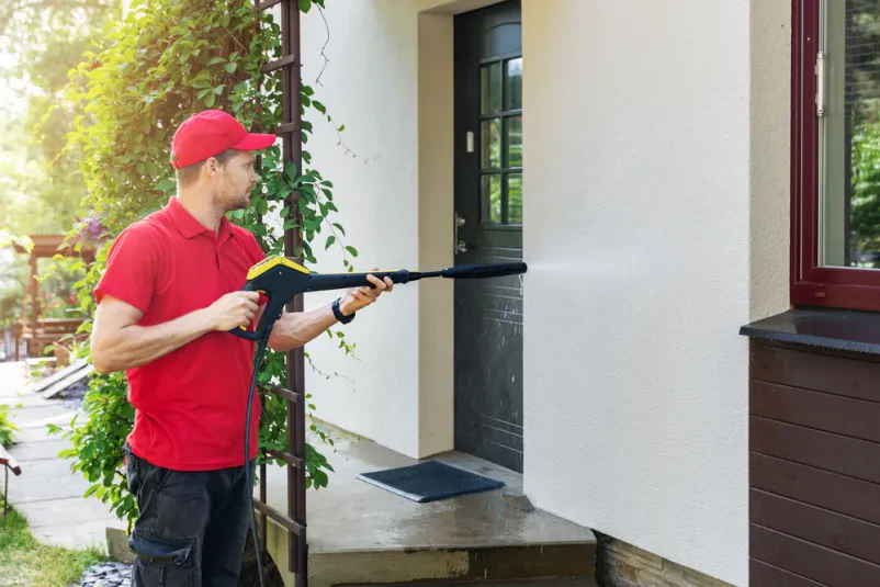 Delavec čisti fasado hiše z visokotlačnim pranjem Moški v delovni obleki s strokovnostjo uporablja visokotlačni čistilec za osvežitev fasade, odstranjujoč umazanijo in madeže.