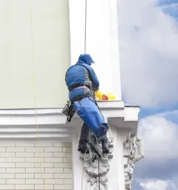 Izkušen delavec pleza po zunanji steni stare stavbe, kar nakazuje na specializirano vzdrževanje ali obnovo fasade, kjer je potreben dostop na višini.