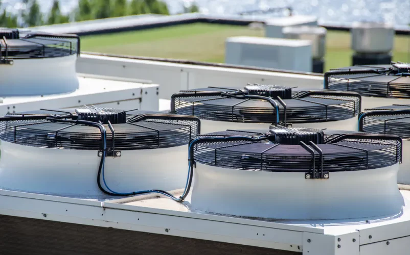 Komercialne HVAC enote na strehi poslovne stavbe Več komercialnih HVAC enot na strehi poslovne stavbe, ki vključujejo hladilne ventilatorje in napredno tehnologijo za nadzor temperature.