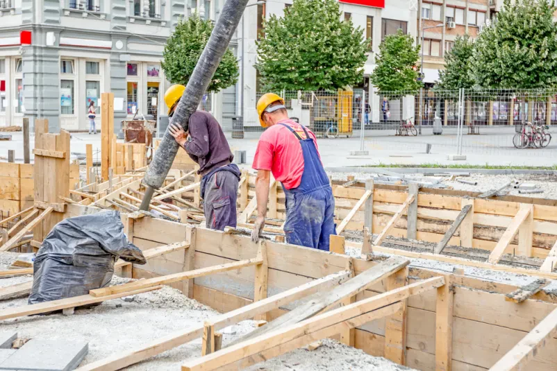 Delavci vlivajo beton v leseni opaž na gradbišču Gradbeni delavci s pomočjo cevi usmerjajo svež beton v leseni opaž, skrbno pripravljen za vlivanje. Prikaz delo z lesom pri betoniranju.