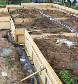 Širši pogled na gradbišče, kjer so pripravljeni leseni opaži in kovinske palice, povezani z žico, kot priprava za vlivanje temeljev.