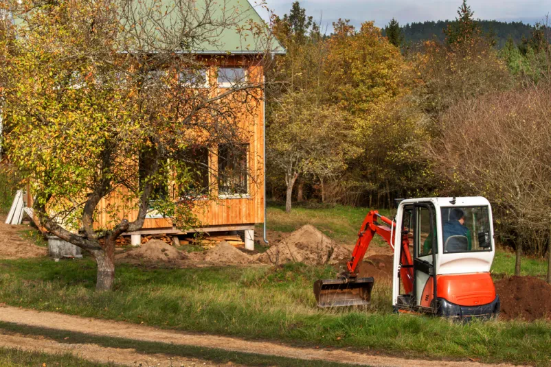 Mini bager ureja teren ob hiši Majhen bager z žlico natančno ravna teren ob novogradnji ekološke hiše, v ozadju pa se razprostira zeleno rastlinje.