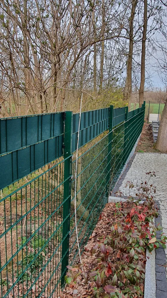 Zasebna ograja iz mrežastih panelov z letvicami Moderna zelena žična mreža z vgrajenimi lesenimi lamelami za zasebnost, ki poteka ob tlakovani kamniti poti. Okolica vključuje vrtne gredice in jesenske liste.